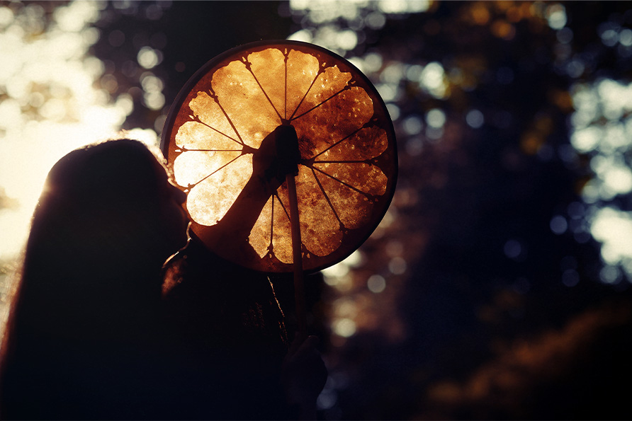 header Cultural History of Shamanic Healing The Stories Behind Sananga Eyedrops Tribal Rapé and Shamanic Tools A person holding a drum in a sunlit forest, with the drum glowing as the light passes through it, creating a silhouette effect.